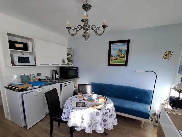 La pièce de vie : la table ronde et sa nappe fleurie, le lit banquette bleu, le lustre et la kitchenette