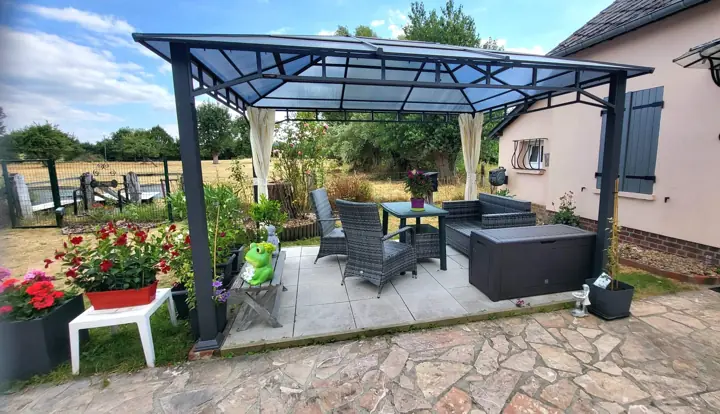 La tonnelle et son salon de jardin sur la terrasse dallée, devant les champs